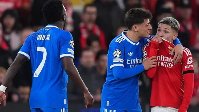 Gianluca Prestianni, con uniforme rojo, se cubre la boca con la camisa mientras se dirige a Vinicius Jr, con uniforme azul, quien está al frente de él. A Prestianni lo abraza el compañero de equipo de Vinicius Jr., Arda Güler