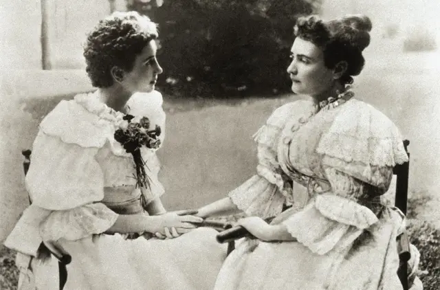 Las dos mujeres, sentadas en un jardín, con vestidos claros de mangas embombadas, con encajes y lazos. Se miran a los ojos y tienen sus manos juntas, pues era su forma de comunicación.