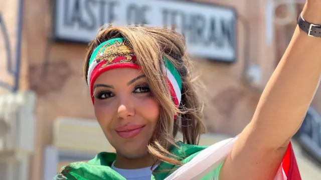 Una mujer iraní-estadounidense posa haciendo el gesto de la victoria con su mano izquierda, con la bandera previa a la Revolución Islámica de Irán con el león y el sol en el centro, que lice también en una cinta en la frente, afuera del restaurante Taste of Tehran en Los Ángeles, California, Estados Unidos, el 15 de junio de 2026. (Foto: Leire Ventas para BBC News Mundo).