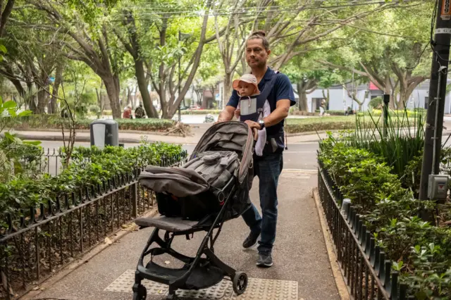 Un padre primerizo carga a su pequeño hijo en un portabebés y empuja un chochecito a través de un parque con árboles y arbustos