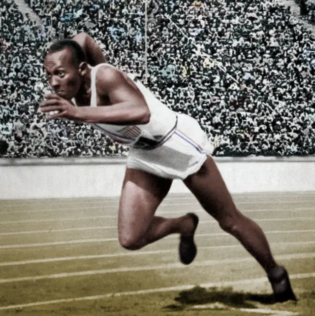 Fotografía en blanco y negro coloreada muestra a Owens con un píe en la pista y otro doblado, inclinado hacia adelante, vestido con camiseta sin mangas y short blancos con rayas delgadas azules y rojas, y zapatillas negras. En el fondo, una multitud en la gradería 