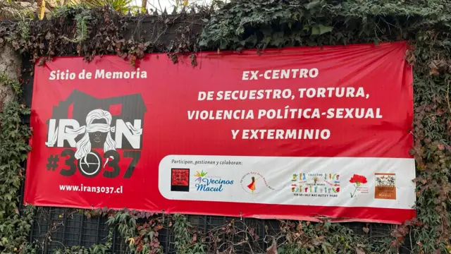 Un primer plano de una pancarta roja situada fuera de los muros del lugar conmemorativo, anteriormente conocido como «Venda Sexy». La pancarta lleva una inscripción en letras blancas y negritas que reza: «antiguo centro de detención, tortura, violencia político-sexual y exterminio». La pancarta también incluye un dibujo de la casa en negro, el nombre «Irán #3037» y el dibujo de una mujer con los ojos vendados.