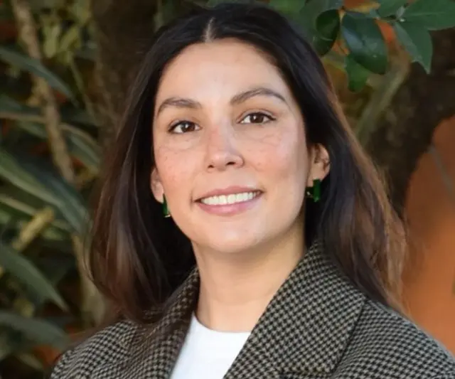 Dra Lozano, una mujer joven, sonriendo, cabello castaño y ojos cafés, vestida con una camiseta blanca y un blazer de cudros negros y taupe. En el fondo se ve una planta.