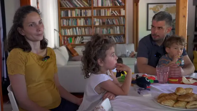 Una mujer y un hombre están sentados alrededor de una mesa, cada uno con dos niños pequeños en su regazo. Sobre la mesa hay pasteles y juguetes. Al fondo se ve una estantería, un sofá y, encima de este, una guitarra.
