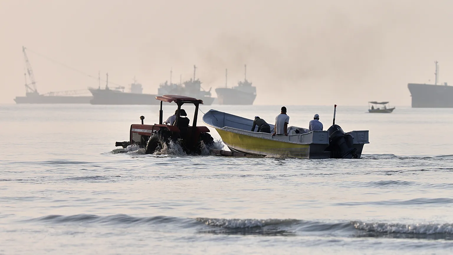 A tractor tows a small boat through shallow water as cargo ships and industrial vessels are anchored in the Strait of Hormuz off Bandar Abbas, Iran, Monday, June 1, 2026. 