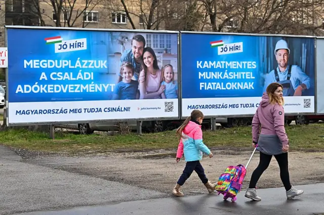 Una madre y su hija caminan por la acera, frente a dos carteles publicitarios. El primero muestra a una familia de cuatro personas con el lema "Duplicamos las ayudas fiscales para familias". El segundo muestra a un trabajador y el lema "Préstamos sin intereses para jóvenes trabajadores".
