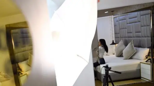 Una chica se prepara para hacer una sesión por webcam en una habitación: se le ve sentada en la cama de espaldas, mientras se arregla el cuello de la camisa antes de salir en vivo. El rostro está desenfocado.