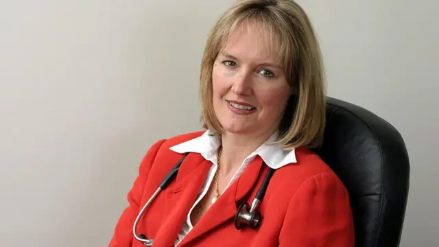 La doctora Helena Teede, una mujer rubia vestida con blusa blanca y chaqueta roja, con un estetoscopio colgando de su cuello, y sentada en una silla negra.