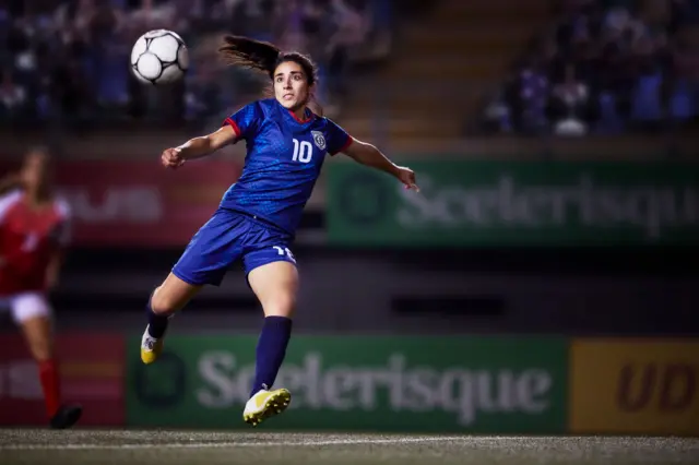 Mujer vestida con equipación deportiva de fútbol, camiseta de manga corta, pantalones cortes, medias altas, todo de color azul, botines de tacos de color amarillo. Está en posicion de golpear un balón que está en el aire. 
