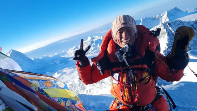 Purnima Shrestha haciendo un gesto de victoria y con un humor jubiloso de pie en una montaña del Himalaya cubierta de nieve.