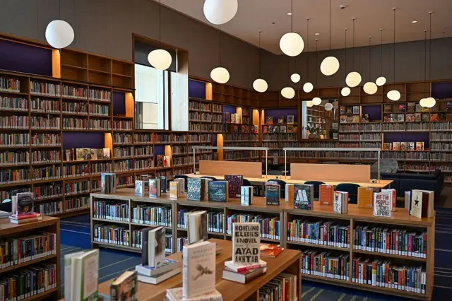 La habitación está repleta de estanterías y libros expuestos en madera clara. Hay una alfombra azul y verde, y del techo cuelgan lámparas esféricas de formas irregulares.
