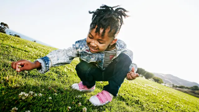 Una niña sonríe mientras está agachada en cuclillas profundas sobre un campo de césped iluminado por el sol. Está buscando pequeñas flores blancas. La niña lleva una chaqueta vaquera, pantalones oscuros y zapatos rosas.