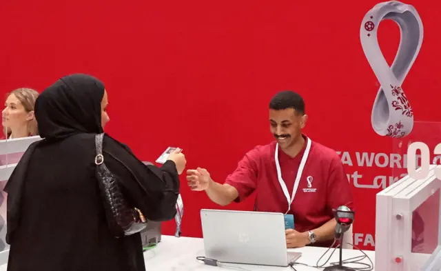 Un hombre sonriente con una camiseta roja ayuda a una mujer vestida de negro. Ella sostiene algo que parece una tarjeta de identificación con un cordón. La pared que hay detrás del hombre es de color rojo brillante.