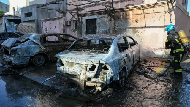 Un bombero echa agua con una manguera en dirección de unos autos quemados por la caída de esquirlas de drones