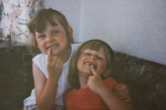 Dos niñas sonriendo a la cámara, ambas señalan los huecos dejados por sus dientes faltantes.