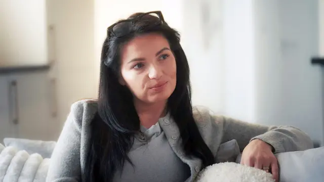 Leanne está sentada en un sofá blanco, mirando a alguien fuera de cámara con expresión seria. Tiene el cabello largo y oscuro, y lleva unas gafas colocadas sobre la cabeza.