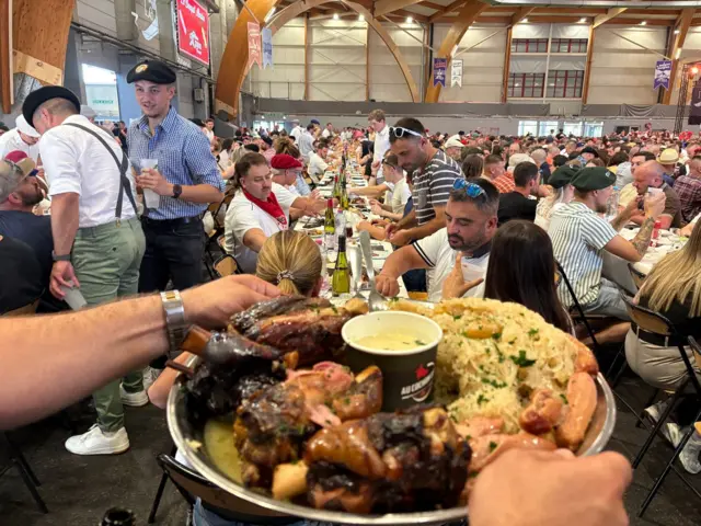 En primer plano de la imagen se ve un plato grande de carne y patatas. Un hombre que lleva un reloj de pulsera metálico lo sostiene sobre una bandeja de plata. Al fondo, cientos de personas están sentadas a largas mesas; hay botellas de vino dispuestas a lo largo del centro de las mismas.