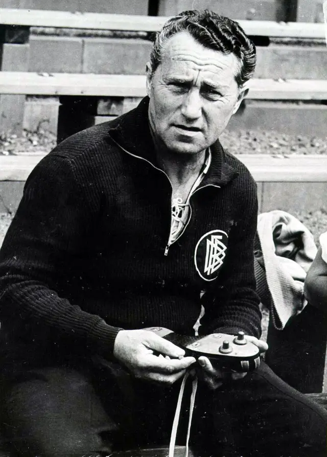 Fotografía en blanco y negro de Adolf Dassler, fundador de la empresa alemana de ropa deportiva Adidas, con ropa oscura y la insignia de la Federación Alemana de Fútbol (DFB) a un lado en el pecho, sosteniendo zapatillas de fútbol con dos tacos y huecos para fijar más. 