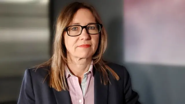 Rachel tiene el cabello hasta los hombros y lleva gafas, una chaqueta oscura y una camisa clara abotonada; se encuentra ante un fondo de interiores.