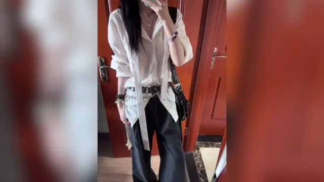 Mujer de pie haciéndose un selfie frente a un espejo con una pared roja en el fondo, vestida con pantalones negros y camisa blanca con flecos largos