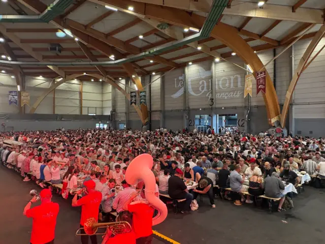 Filas y filas de mesas largas abarrotadas de gente en un gran salón de techos muy altos. En la pared del fondo de la sala están pintadas las palabras "Le Canon Français". En primer plano aparece una banda de música de viento-metal de pie, que incluye una tuba, una trompeta y dos cornetas; sus integrantes visten camisetas y gorras de béisbol rojas.