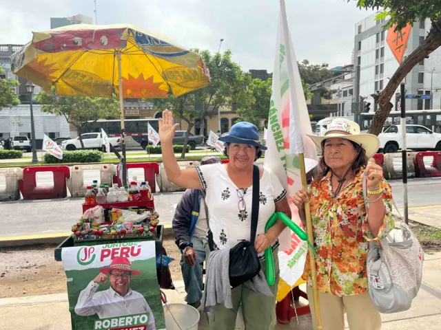 Dos seguidoras del candidato Roberto Sánchez, al lado de un puesto de ventas ambulantes con un letrero de Sánchez Presidente