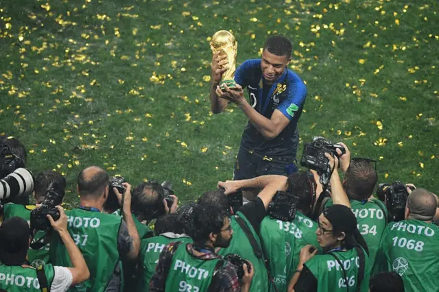 Mbappé sostiene la copa del mundo mientras los fotógrafos disparan sus cámaras.