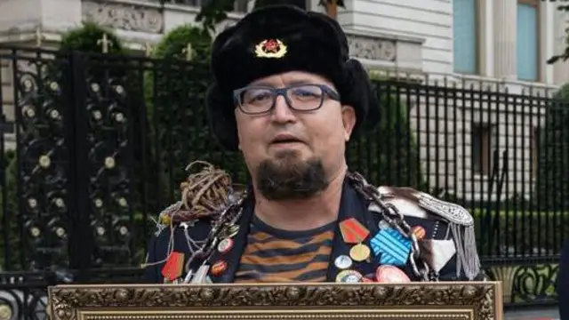 Un hombre que lleva un gorro ruso de estilo antiguo con una insignia soviética sostiene la parte superior de un cuadro enmarcado y viste una chaqueta repleta de medallas.