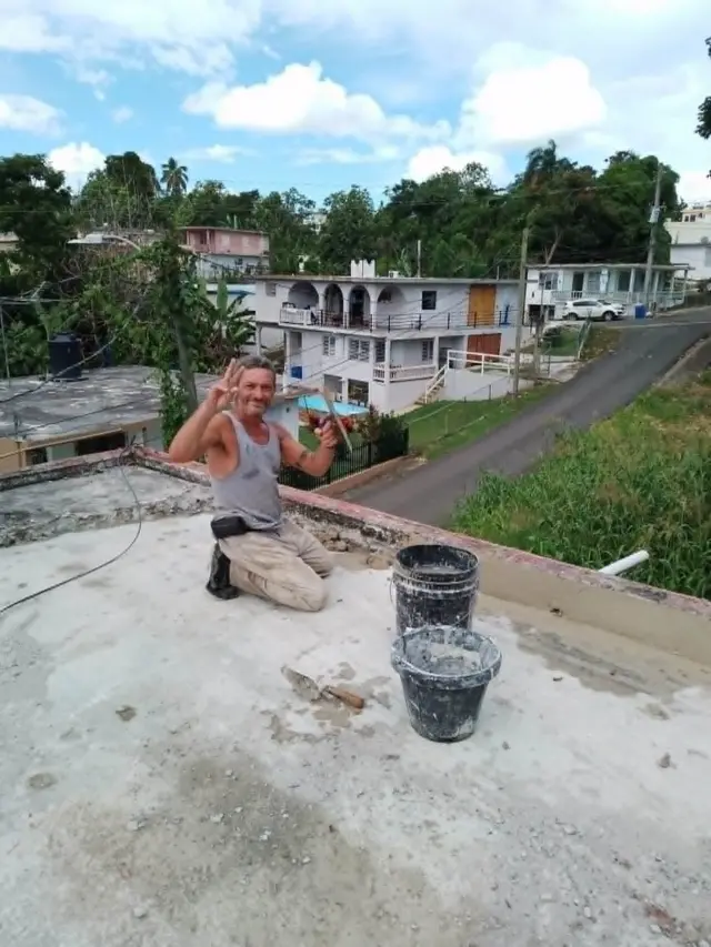 Lázaro Romero León saluda a cámara haciendo el gesto de la victoria con la mano derecha y una paleta en la izquierda, sobre el techo de una vivienda, junto a unos cubos con cemento, en Puerto Rico.