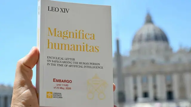 Primer plano de una mano sosteniendo un ejemplar de Magnifica humanitas. En la portada aparece el título de la encíclica, además del embargo que la afecta, en letras rojas. Al fondo se aprecia un cielo azul y lo que parece ser la basílica de San Pedro en Roma.