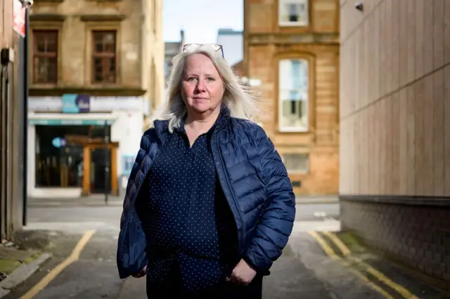 Christine Goodall, cirujana oral, lleva una chaqueta azul y una camisa azul de pepas blancas. Tiene las manos en los bolsillos y mira directo a la cámara.
