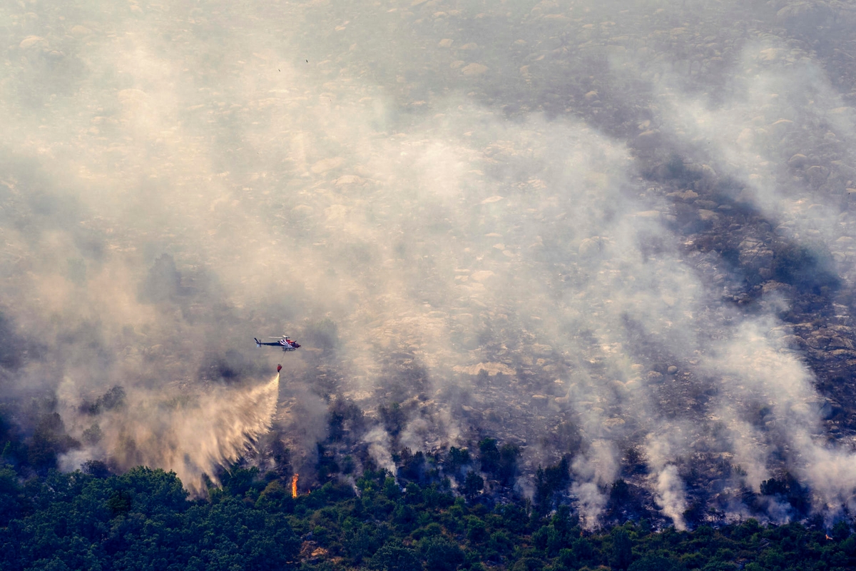 Un helicóptero arroja agua sobre una zona de incendio forestal con llamas, humo, vegetación verde y suelo quemado