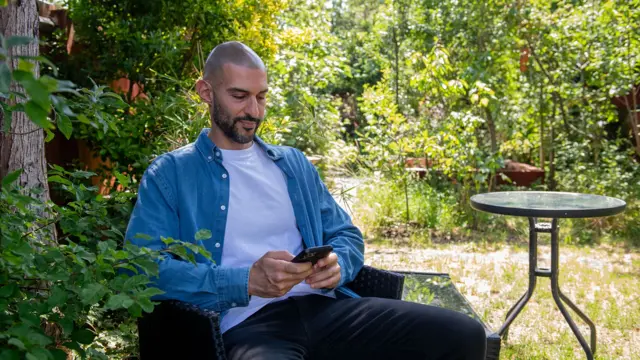 Un hombre con bigote y barba oscuros con camisa azul, camiseta blanca y pantalones negros, sentado en un jardín, mirando su teléfono móvil.