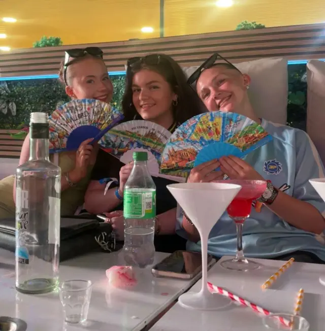 Tres mujeres jóvenes sentadas en un bar, sosteniendo abanicos de papel.