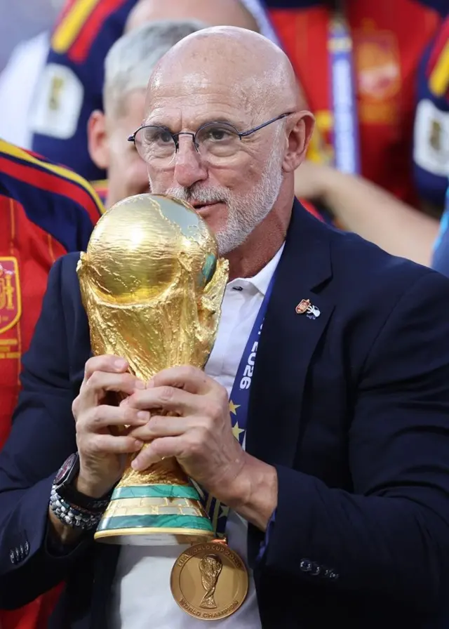 Luis de la Fuente, con lentes, camisa blanca y chaqueta azul oscura, carga el trofeo del Mundial con las dos manos. Está rodeado de futbolistas españoles. En su cuello, cuelga la medalla de oro