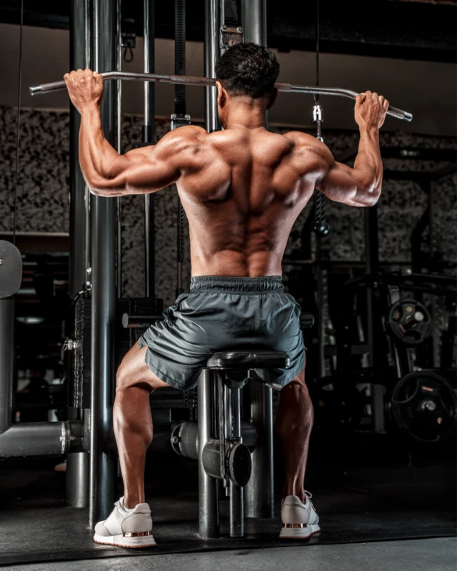 En la foto, Jamal aparece de espaldas, sentado en una máquina de polea alta lateral. Lleva pantalones cortos, no lleva camiseta y tiene una musculatura muy desarrollada.