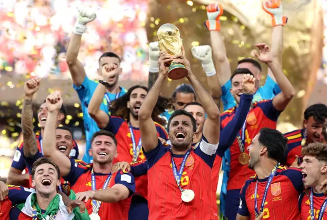 Rodri, en el centro levanta la copa del mundo con las dos manos. Está rodeado de sus compañeros de la selección española que están felices. Algunos levantan los brazos. Otros se abrazan. Visten el uniforme y llevan colgadas las medallas de oro