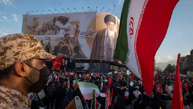 Un agente de seguridad iraní entre la multitud frente a una valla publicitaria que muestra al difunto líder supremo en su funeral.
