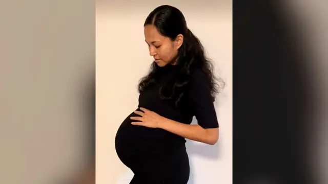 Jenitar Na'amoana aparece en una fotografía durante su embarazo. Tiene el cabello largo y oscuro y lleva un vestido negro de manga corta. Mira hacia abajo y toca su prominente barriga.