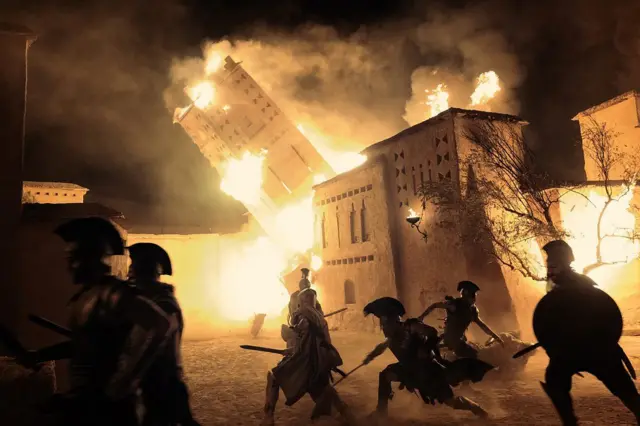 Edificios envueltos en llamas durante una escena nocturna, mientras varias personas con escudos y lanzas corren en primer plano entre humo y fuego.