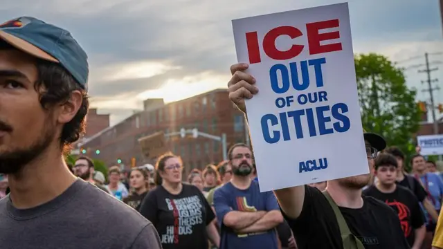 Fotografía de una multitud con un cielo gris de fondo. Una persona lleva una pancarta que dice: "¡Fuera ICE de nuestras ciudades! ACLU". No se ve el rostro de quien la porta. La multitud es mixta, con gente joven y mayor, y un joven con gorra destaca a la izquierda de la imagen, cuyo rostro también está parcialmente oculto.