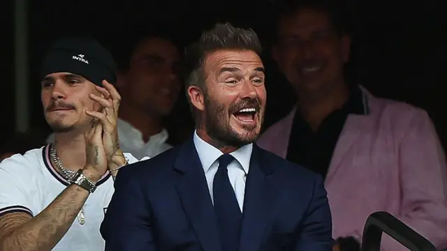 El exfutbolista inglés David Beckham sonríe durante el partido de cuartos de final del Mundial de Fútbol de 2026 entre Noruega e Inglaterra en el Miami Stadium de Miami Gardens el 11 de julio. Viste un traje azul, camisa blanca y corbata azul.