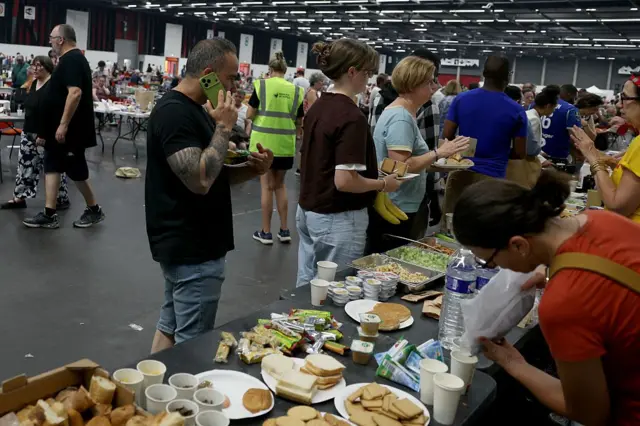 En primer plano, a la derecha, una voluntaria vierte algo en un vaso, en una mesa alargada llena de comida para repartir. Al otro lado, a la izquierda, varias personas hacen fila con platos en la mano para recoger comida. Al fondo de este centro de refugio improvisado hay mucha más gente y mesas. 