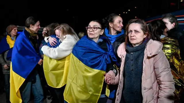 Varias mujeres envueltas en banderas ucranianas azules y amarillas. Dos mujeres se abrazan al fondo y otras sonríen. Liudmyla mira hacia su derecha mientras sostiene la mano de una mujer con abrigo rosa situada a su izquierda.