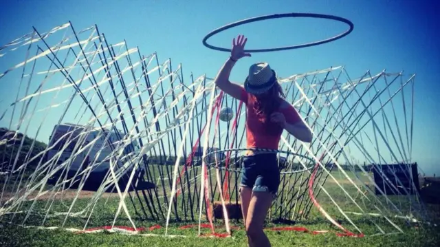 Lowri baila con un aro de hula-hula alrededor de la cintura. Lleva una blusa roja de manga corta y pantalones cortos vaqueros, y se encuentra en un campo con una instalación artística cubierta de serpentinas detrás de ella.