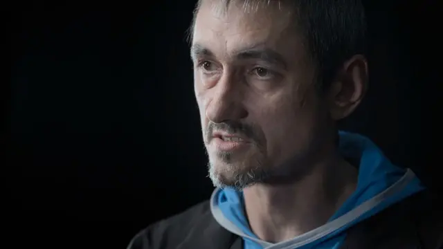 Retrato de Oleksii Sivak, quien lleva barba corta y bigote, mira hacia la izquierda de la cámara y viste una sudadera con capucha azul claro debajo de una chaqueta negra.