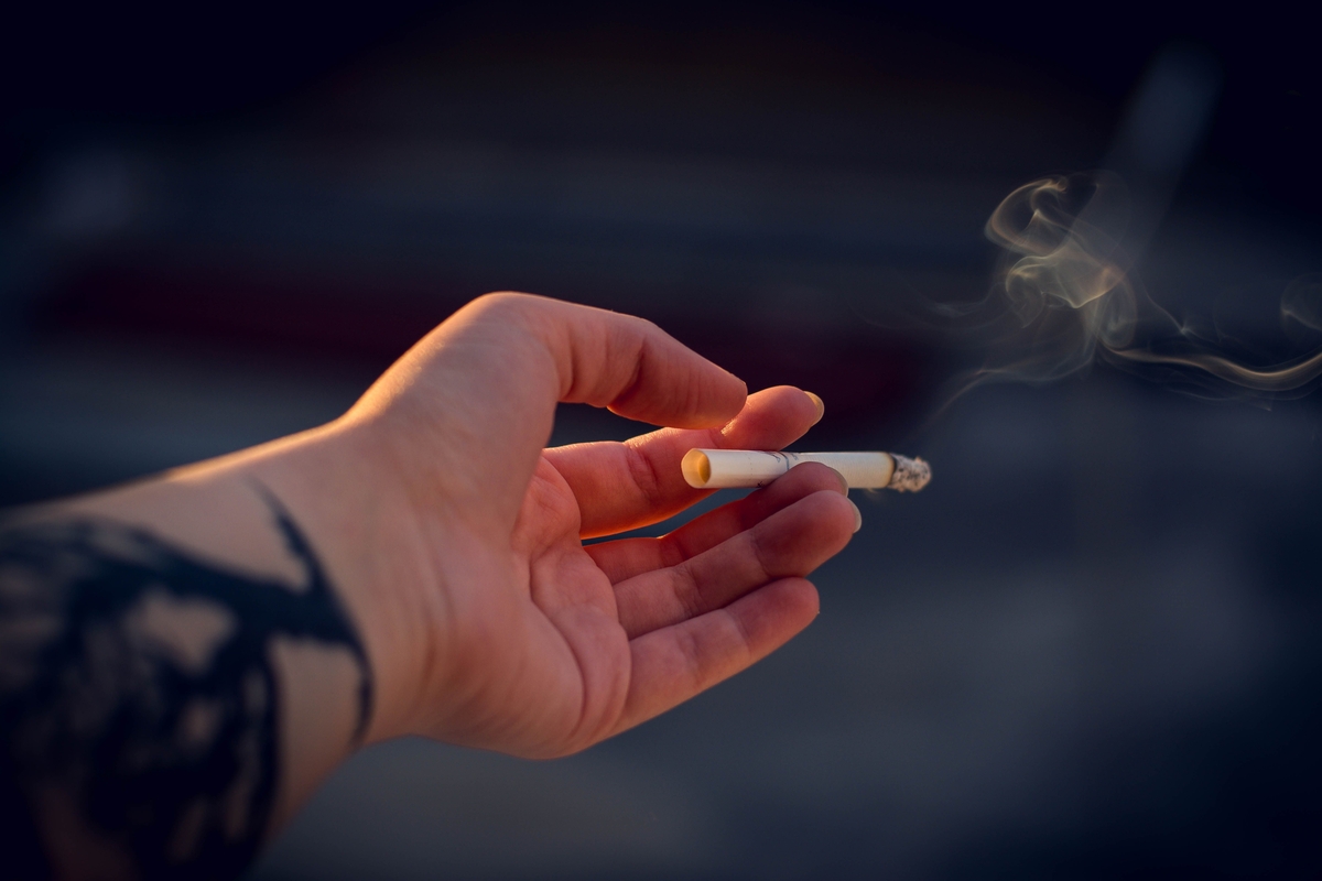 Mano con tatuaje en el antebrazo sujeta un cigarrillo encendido con filtro naranja y ceniza, del que se eleva humo. Fondo oscuro desenfocado