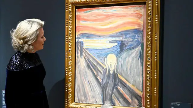 Un cuadro enmarcado que muestra a una persona gritando contra un cielo rojo brillante se exhibe en una galería, con un visitante de pie junto a él contemplando la obra de arte.