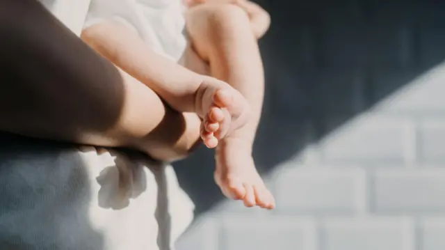 Primer plano de los pies de un bebé. El bebé es sostenido por un adulto que lleva una camisa blanca, de pie frente a una pared de ladrillos grises.