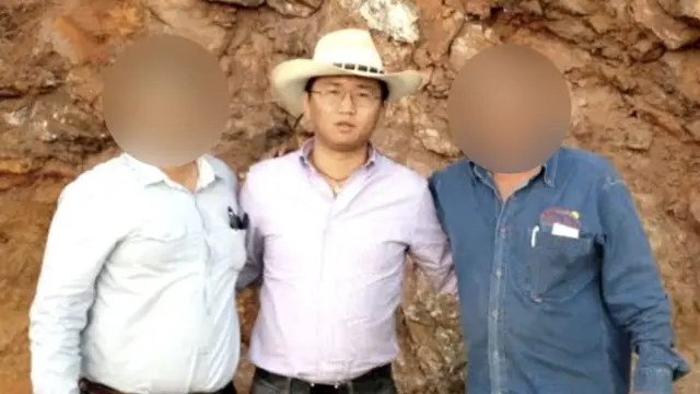 Un hombre lleva un sombrero blanco estilo vaquero y una camisa lila metida por dentro del pantalón. Usa gafas y un collar, y mira a la cámara. A su izquierda hay un hombre y a su derecha otro; los rostros de ambos aparecen borrosos para ocultar sus identidades. 